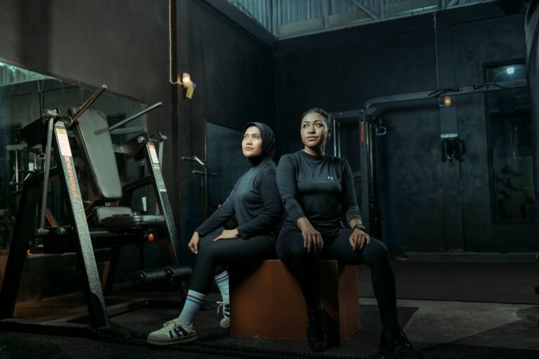 Two women in athletic wear sitting on a box with gym equipment in the background.